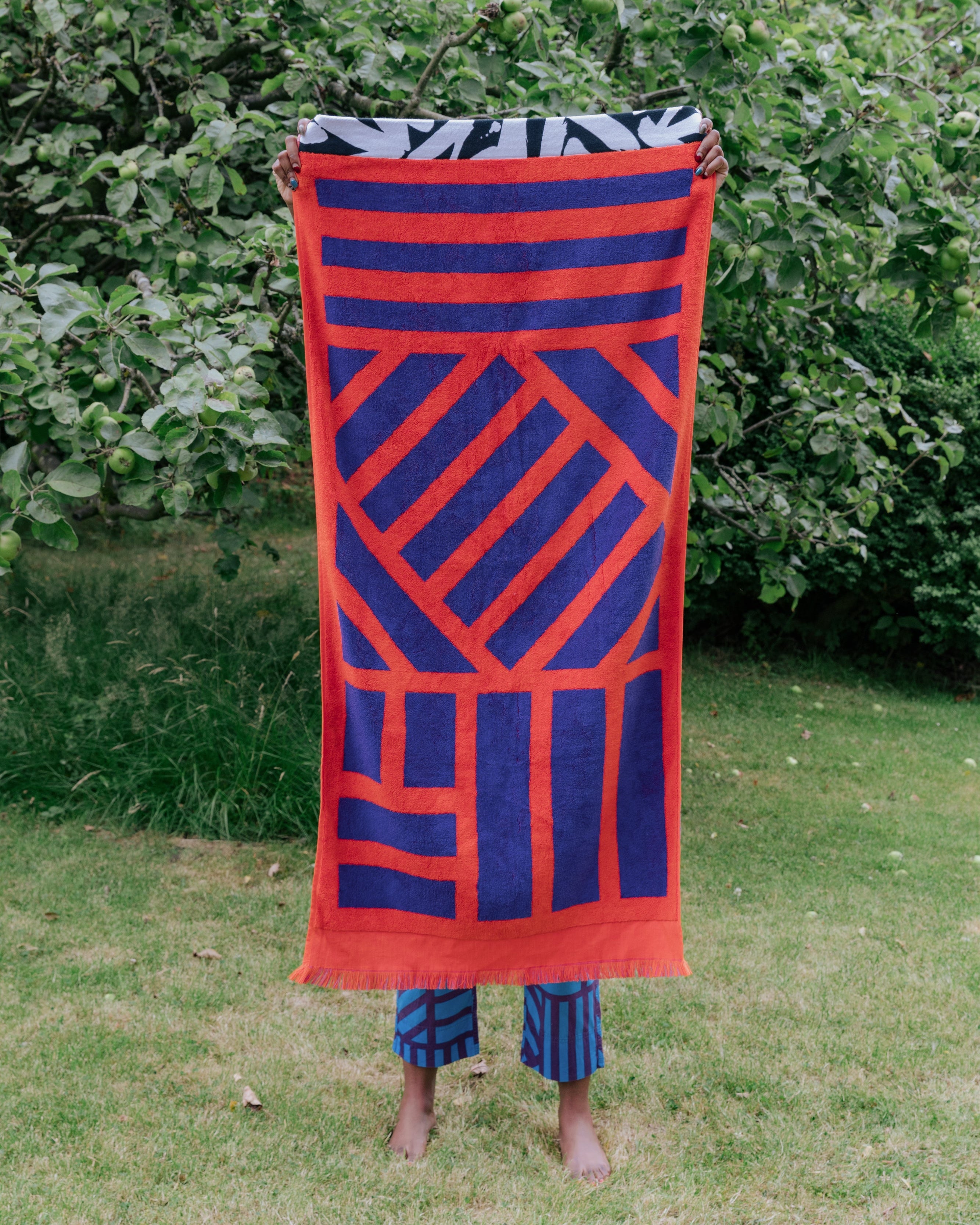 A person standing outdoors on grass in front of leafy green trees holds up a large towel with a bold geometric pattern in red and dark blue, with patterned stripe pants visible below.