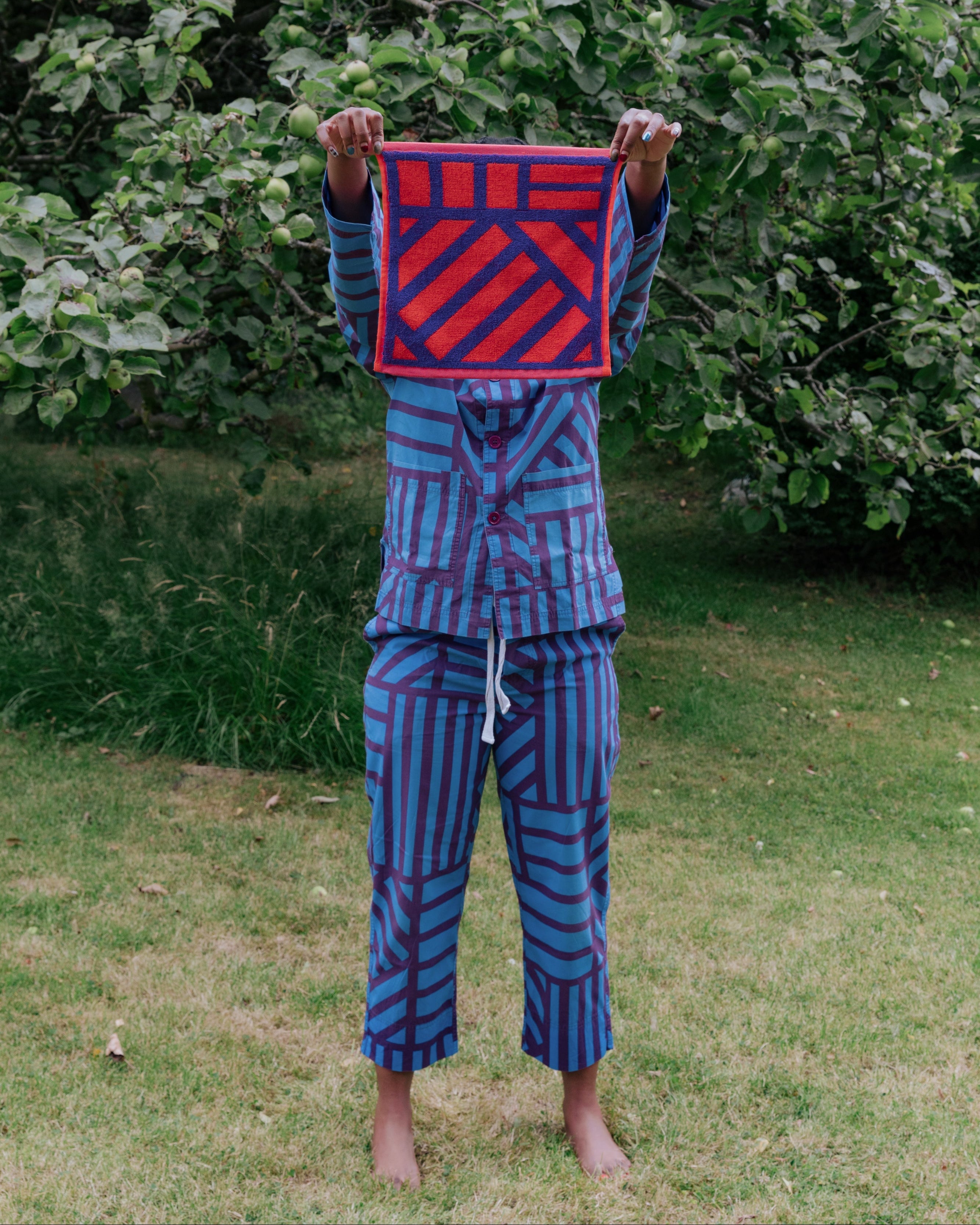 A person standing barefoot in a grassy outdoor area with leafy trees behind them, holding up a facecloth in front of their face. The fabric has a red and purple geometric striped design, and the person is wearing matching blue and purple striped pyjamas