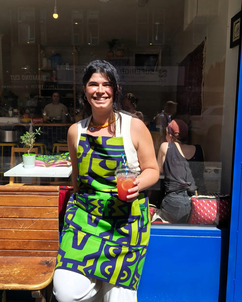 A woman smiling and holding a drink in front of a storefront, wearing a sleeveless apron with a bold green, black, and white geometric pattern.